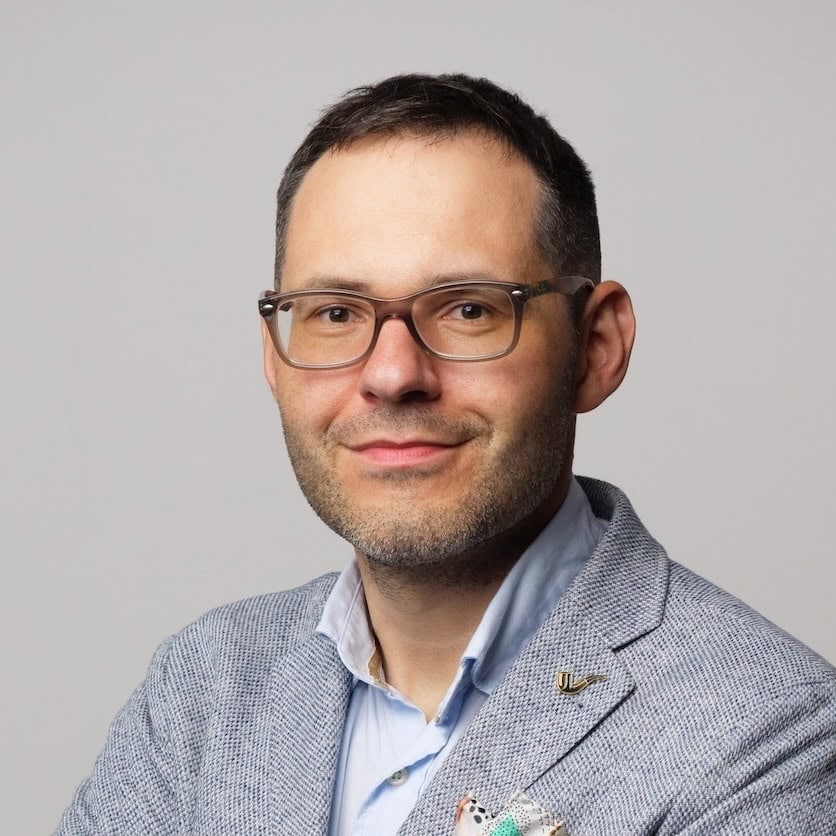 A man with short dark hair and glasses, wearing a light blue collared shirt and a grey textured blazer, smiles softly at the camera against a plain light grey background.