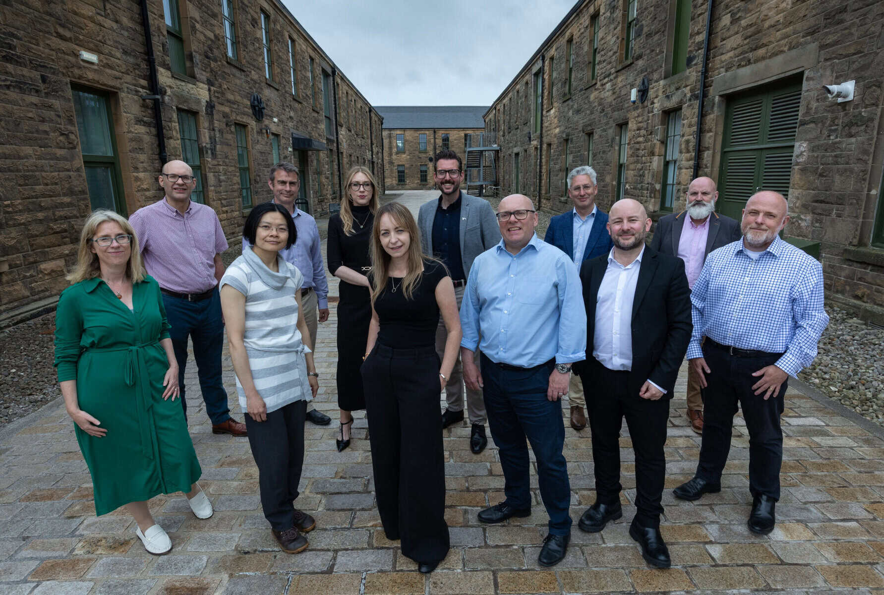 A group of twelve adults, dressed in business casual and formal attire, stand together smiling in a courtyard between two stone buildings under a cloudy sky.