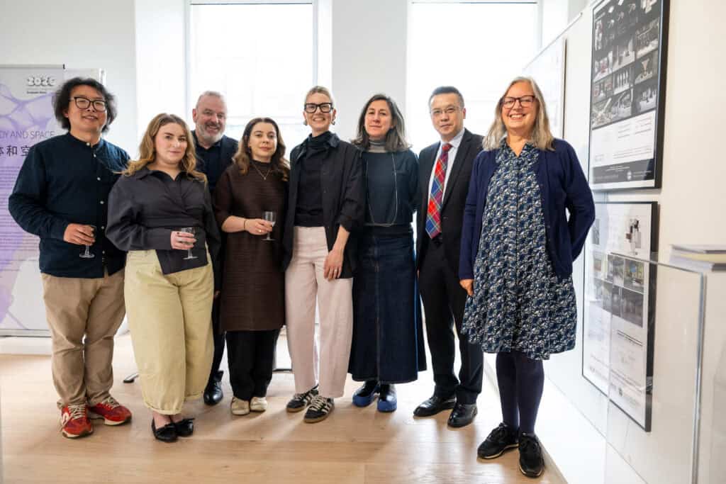 A group of eight people, dressed in smart-casual and business attire, stand smiling together in a well-lit gallery or office space with art and exhibition displays on the walls.