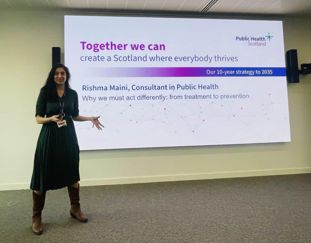 A woman in a dark dress stands and gestures in front of a large screen displaying a Public Health Scotland presentation titled “Together we can create a Scotland where everybody thrives”.