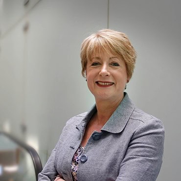 A smiling woman with short blonde hair, wearing a light grey blazer over a patterned top, stands with arms crossed in a modern, softly lit indoor setting.
