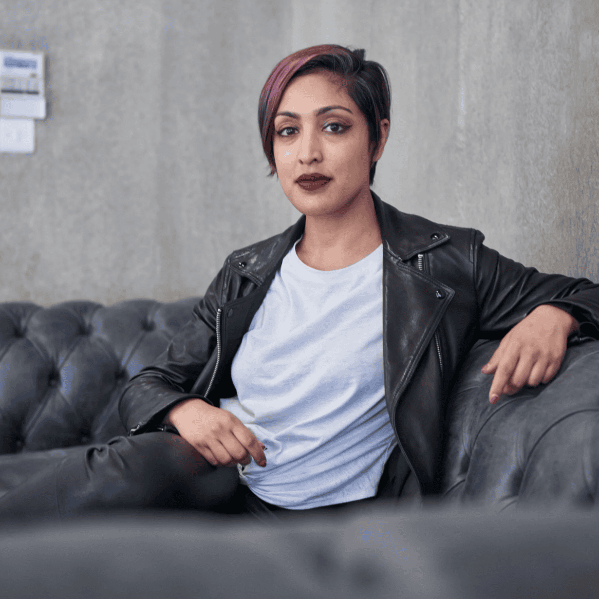 A person with short purple hair sits confidently on a grey tufted sofa, wearing a black leather jacket, white T-shirt, and black trousers, against a concrete wall background.