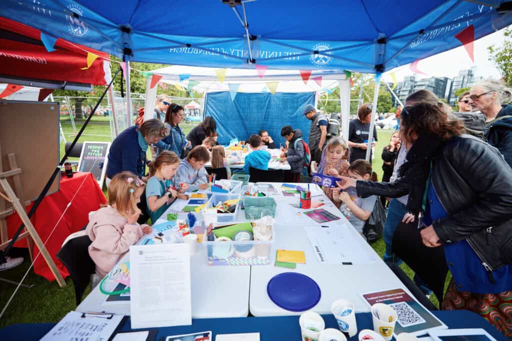 Children and adults participate in arts and crafts activities under a blue marquee at an outdoor event, with colourful bunting overhead and various art supplies on the tables.