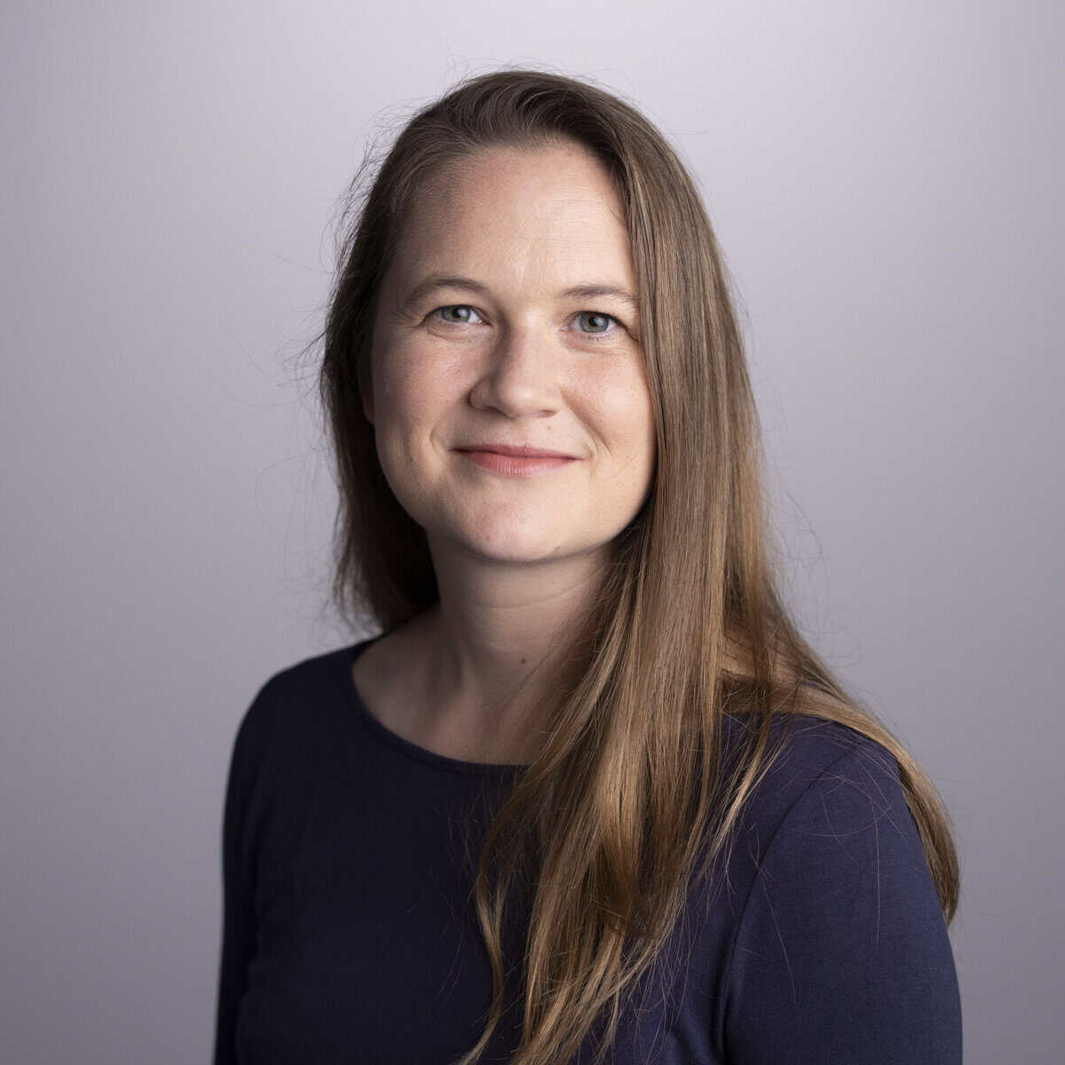 A woman with long brown hair, wearing a dark blue top, smiles softly at the camera against a plain light grey background.