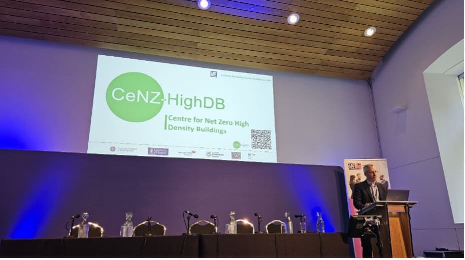 A speaker stands at a lectern in a conference room with a wooden ceiling. A large screen displays "CeNZ-HighDB | Centre for Net Zero High Density Buildings" above an empty panel table with microphones and water jugs.