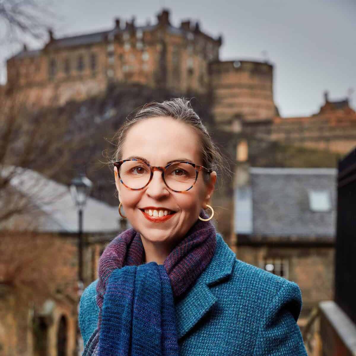 A woman in glasses and a blue coat with a blue scarf smiles outdoors, with an old stone castle on a hill in the background on a cloudy day.