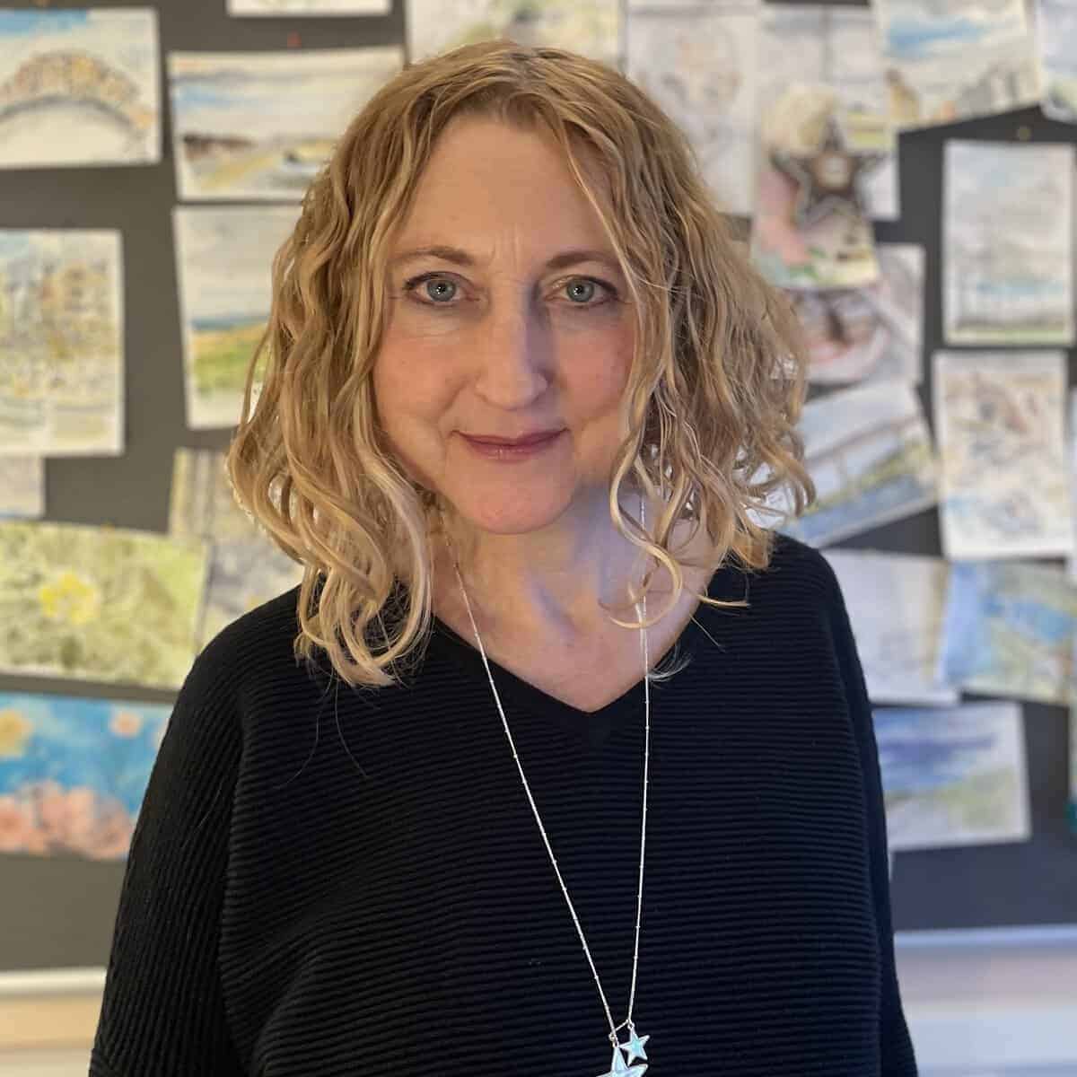A woman with wavy blonde hair, wearing a black top and a long necklace with a star pendant, stands in front of a wall covered with colourful drawings and art.