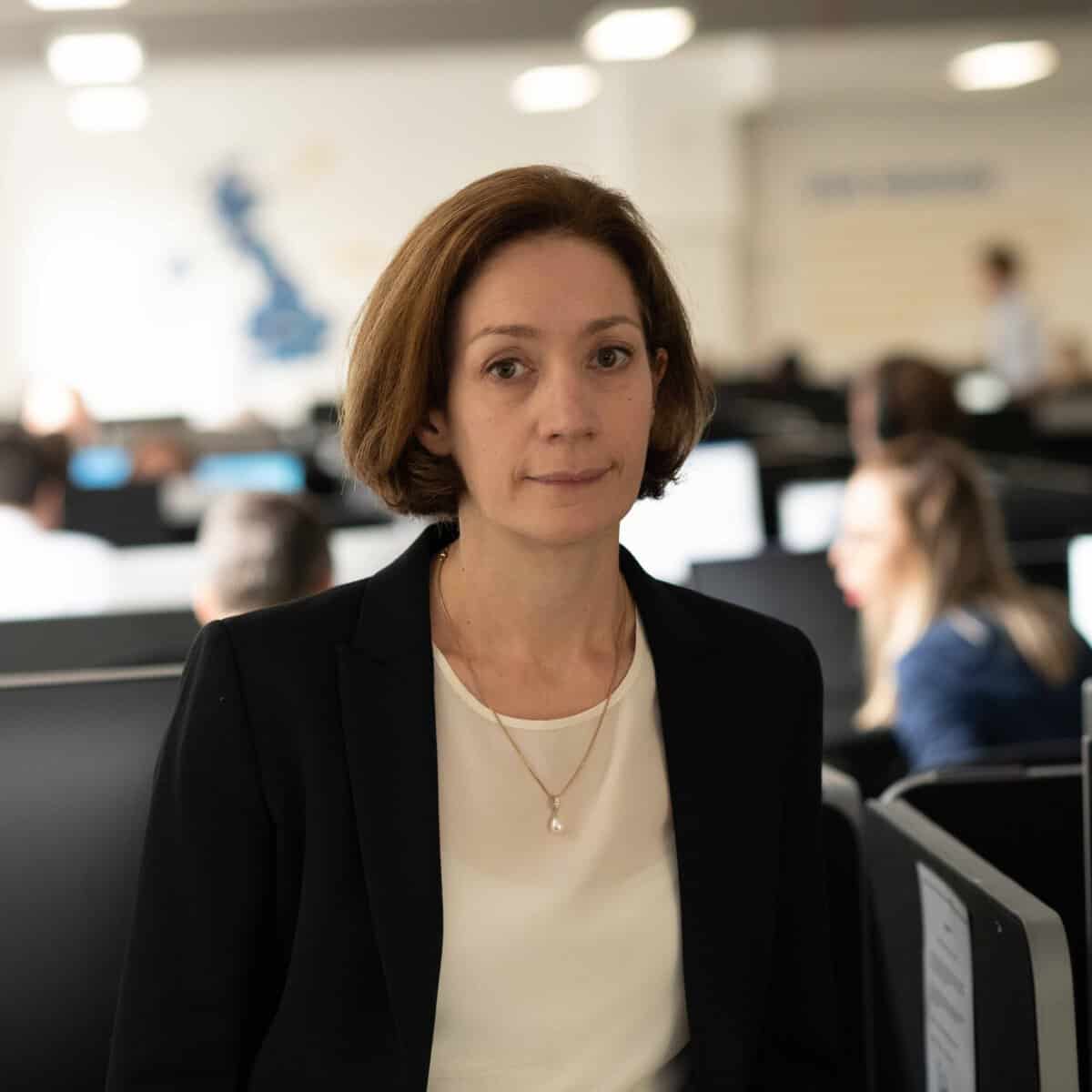 A woman with short brown hair, wearing a black blazer and white top, stands in an office with several people working at computers in the background. The setting appears professional and modern.