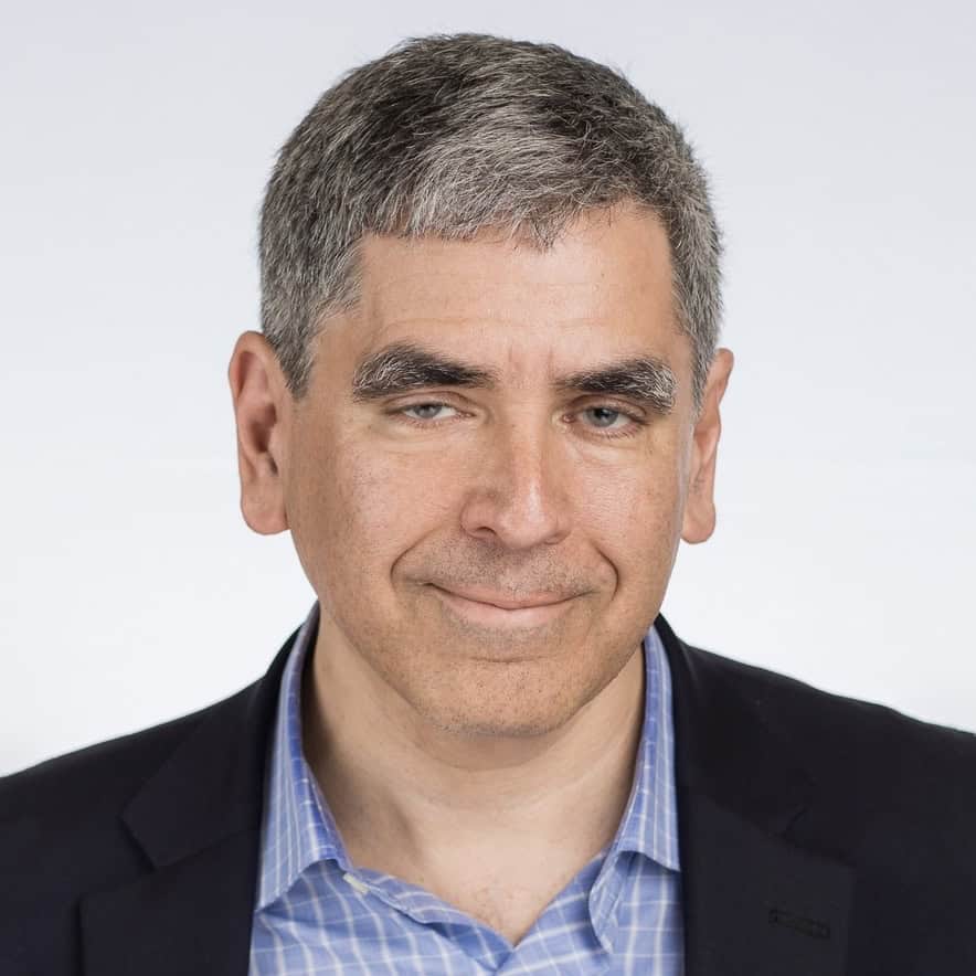 A middle-aged man with short grey hair wearing a checked blue shirt and dark blazer, smiling slightly against a plain white background.