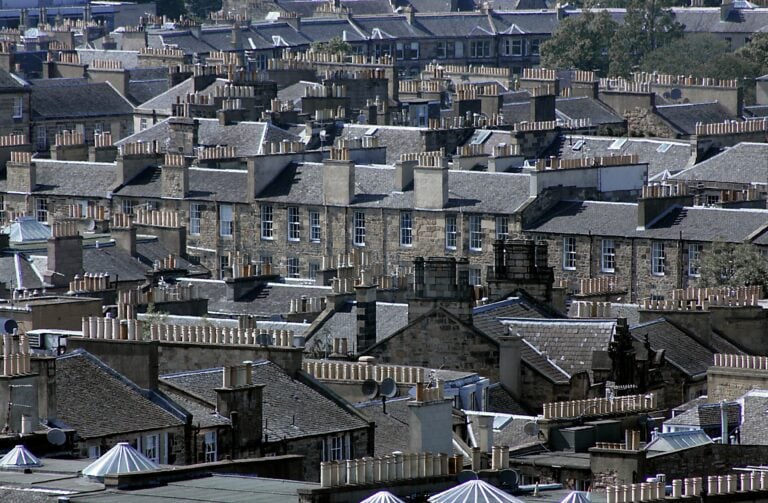 A dense cluster of old stone residential buildings with numerous chimney stacks on their rooftops, seen from above in a tightly packed urban neighbourhood.