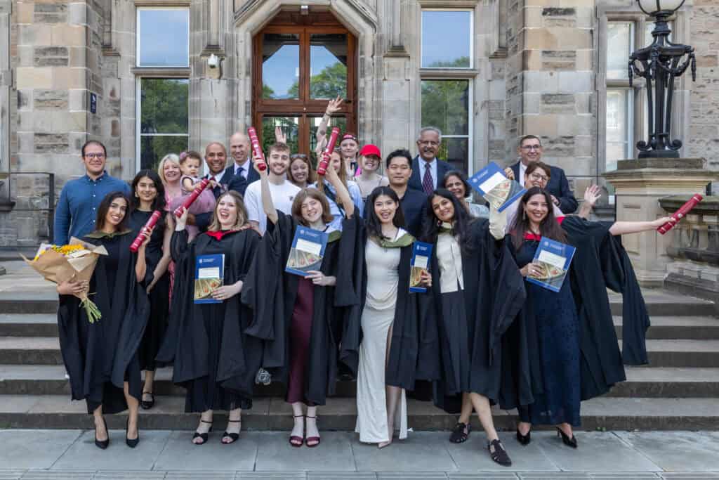 A group of graduates in black gowns stand on stone steps, holding certificates, flowers, and smiling. They celebrate joyfully in front of a historic building.