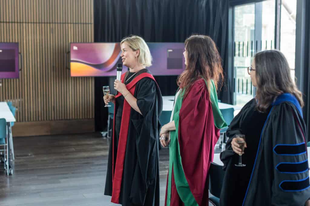 Three women in academic gowns stand indoors, holding champagne glasses. One woman speaks into a microphone whilst the others listen. The background features wooden panels and abstract art on the walls.