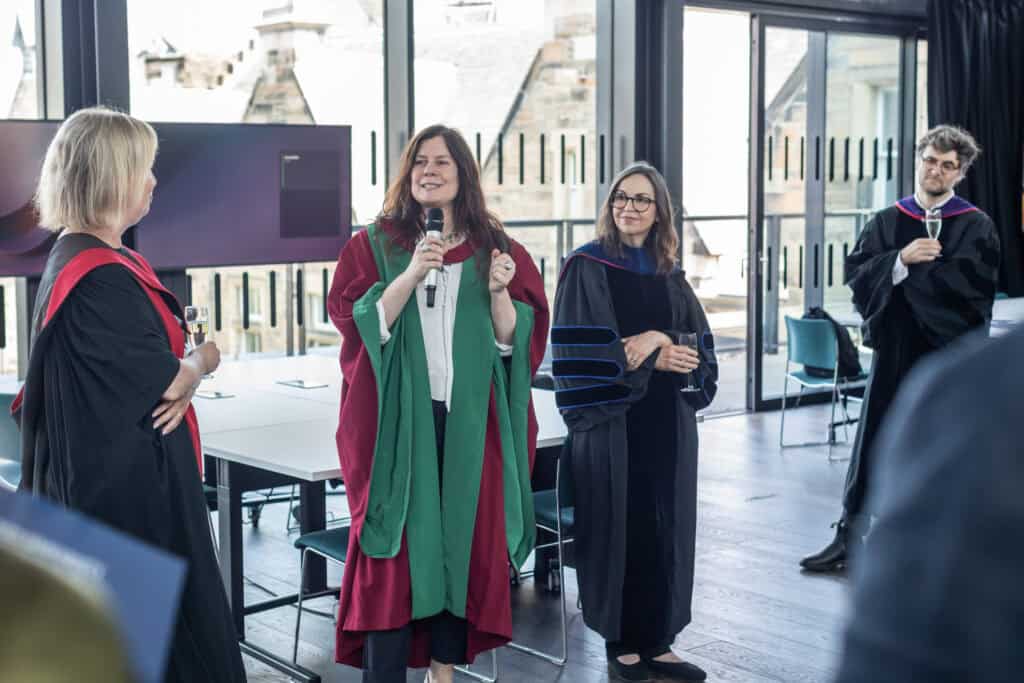 A woman in academic robes speaks into a microphone while others in robes listen and hold drinks in a modern, sunlit room with large windows.