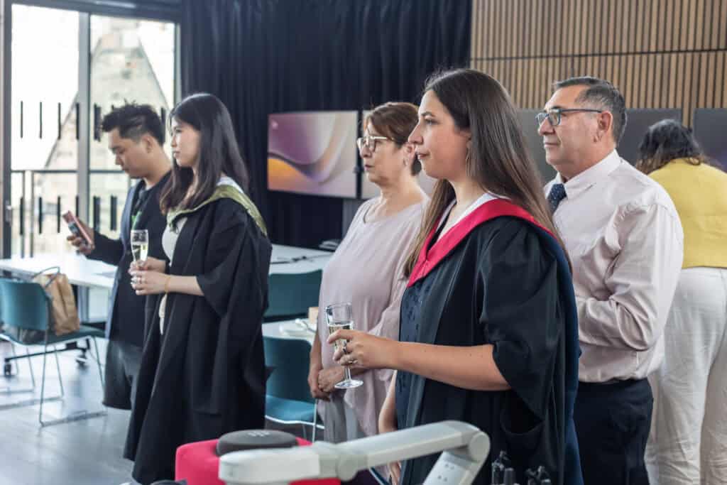 Several people, including two wearing academic gowns, stand indoors at a graduation event, holding drinks and watching something off-camera. Some are dressed formally; tables and modern décor are visible in the background.