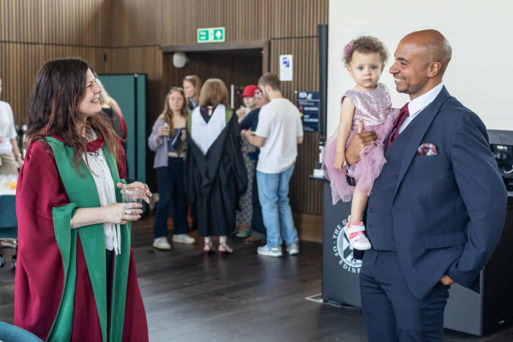 A woman in academic robes smiles at a man in a suit holding a young girl in a pink dress. People are gathered and chatting in the background at an indoor event.