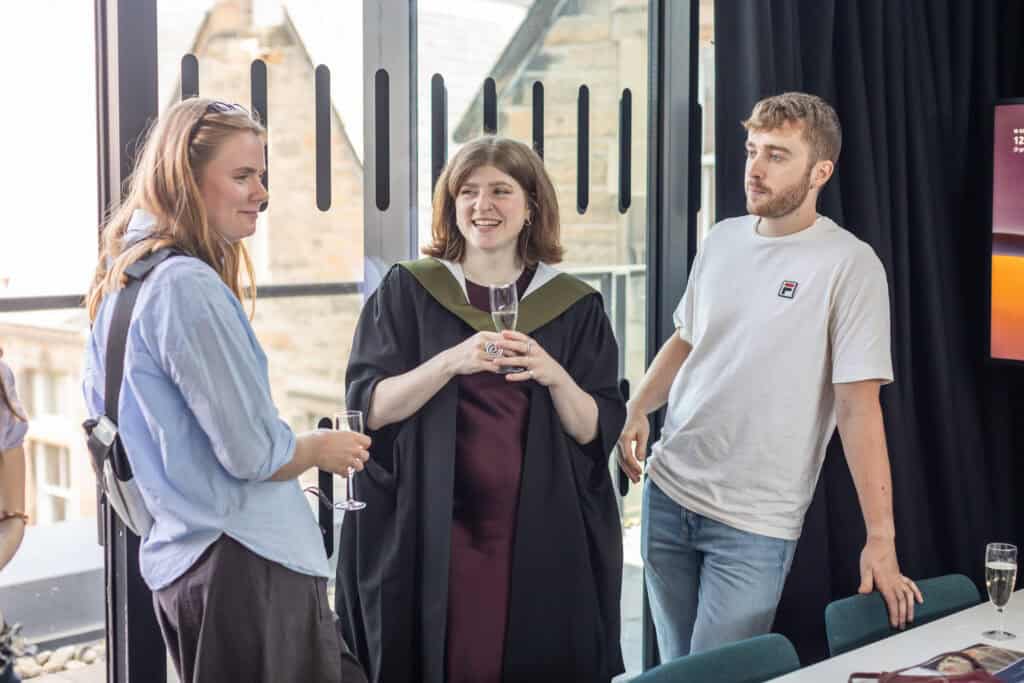 Three young adults converse indoors at a graduation event. One wears a cap and gown holding a champagne glass, while the others, casually dressed, listen nearby. Large windows reveal a stone building outside.