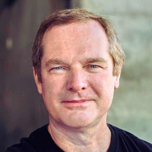 A middle-aged man with short light brown hair and blue eyes wears a black T-shirt and smiles gently whilst looking at the camera. The background is softly blurred in neutral tones.