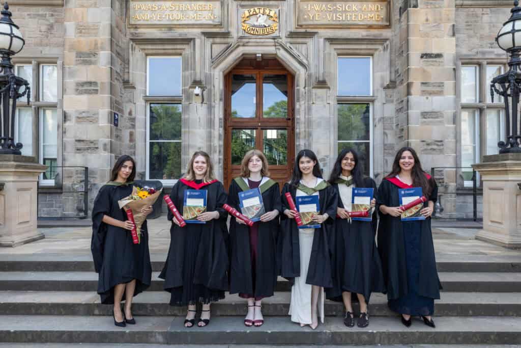 Six graduates in gowns stand on stone steps holding certificates and flowers, smiling in front of a historic building with large windows, lamps, and plaques above the entrance.