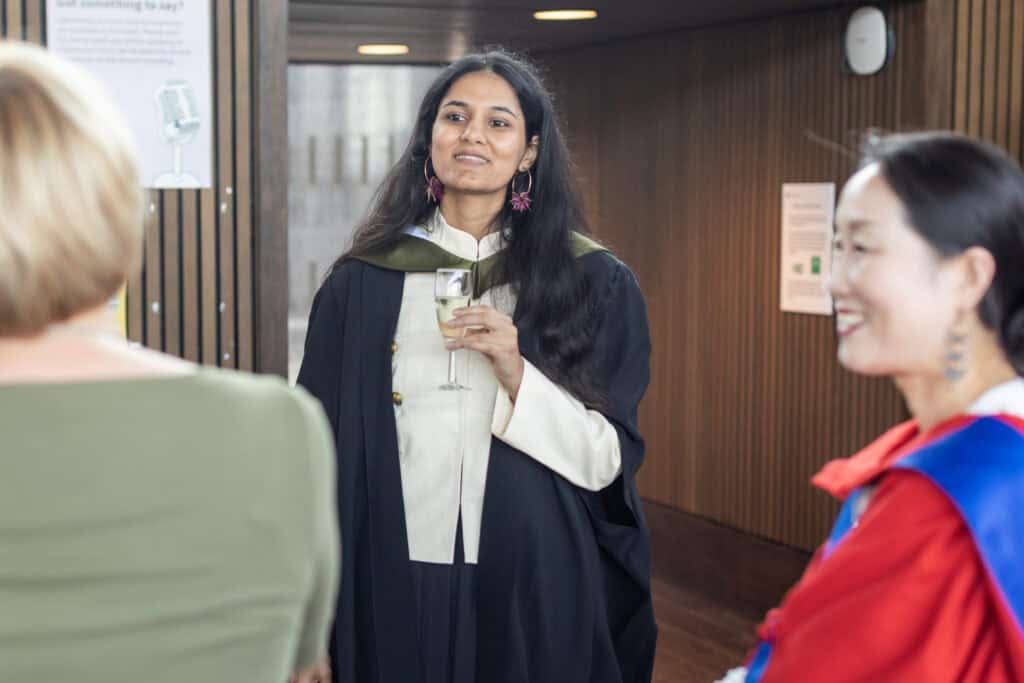 A woman in graduation attire holds a drink and smiles whilst conversing with two others. The setting appears to be indoors, possibly at a ceremony or celebration.