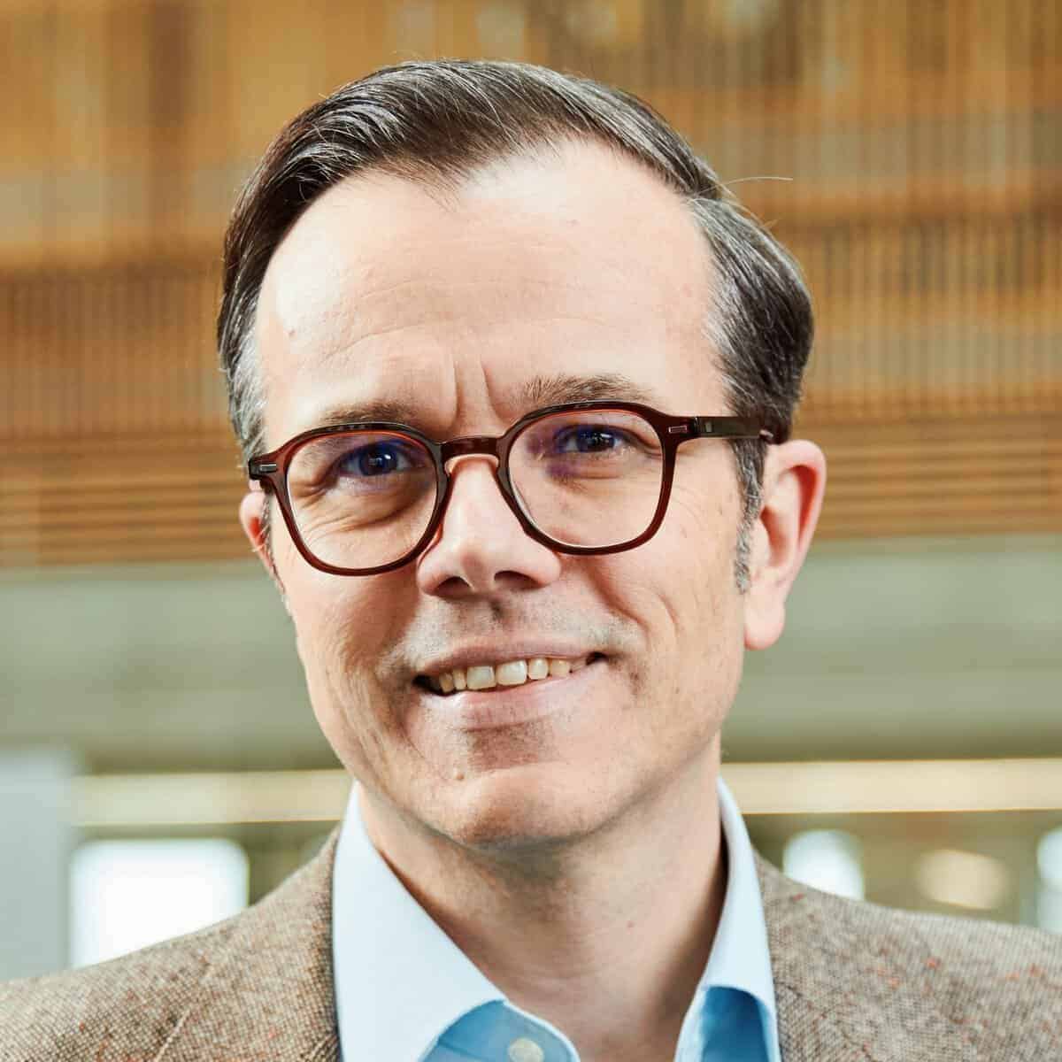 A man with short brown hair, wearing brown glasses, a light blue collared shirt, and a brown tweed jacket, smiles at the camera. The background is blurred with warm-toned wooden slats.