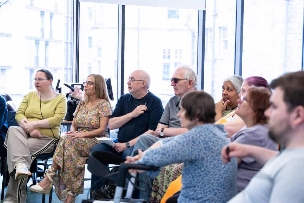 A diverse group of adults sits in a bright room, attentively listening. Some use mobility aids. Large windows in the background let in natural light.