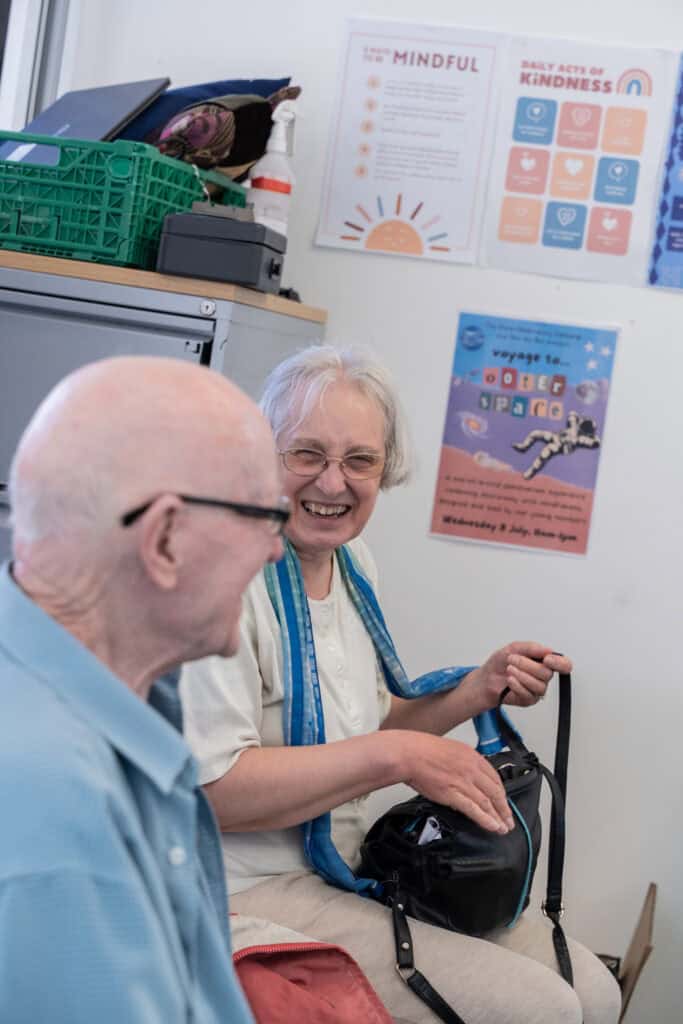 Two older adults sit and laugh together in a bright room. One person holds a black bag and smiles. Colourful posters about kindness and mindfulness are visible on the wall behind them.