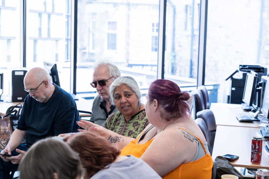 A group of adults sit together in a bright room with large windows, engaged in conversation. Computers are visible in the background.