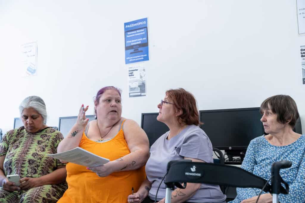 Four women sit in a row in front of computers. One woman in an orange top gestures whilst holding papers, talking to another woman. Signs and posters are visible on the white wall behind them.