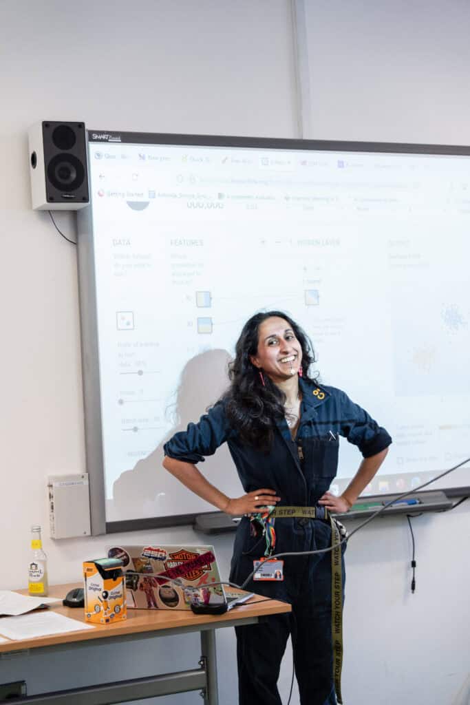 A woman stands smiling in front of a projected presentation screen, hands on hips. A laptop with stickers and a drink are on the desk beside her. She appears to be giving a lecture or presentation in a classroom.