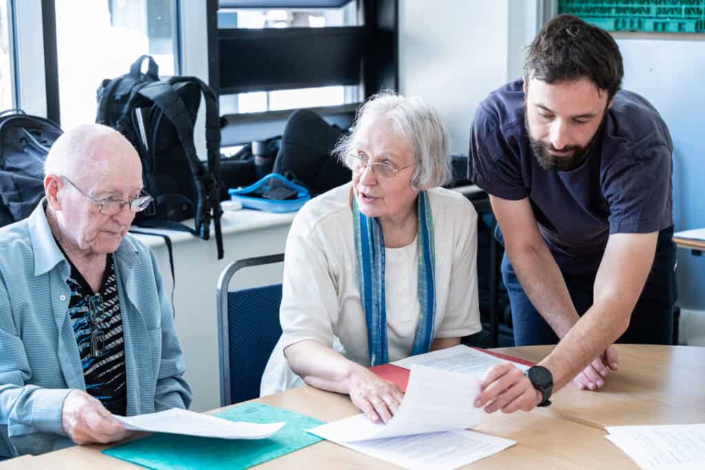 Three people sit at a table with papers, discussing documents. An older man in glasses and an older woman sit whilst a younger man stands, leaning over to help. Rucksacks and windows are visible in the background.