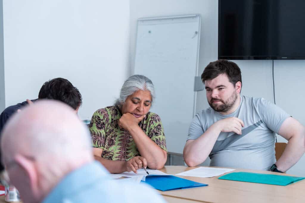 Three people sit round a table, reviewing documents together. A woman in patterned clothing is reading, while a man in a grey T-shirt looks on. A whiteboard and a TV screen are visible in the background.