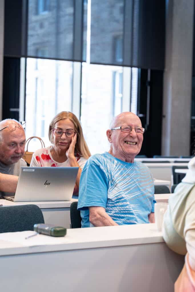An elderly man in a blue shirt smiles whilst sitting in a classroom. Two other adults sit nearby, one with a laptop and another with glasses, all appearing engaged and cheerful.
