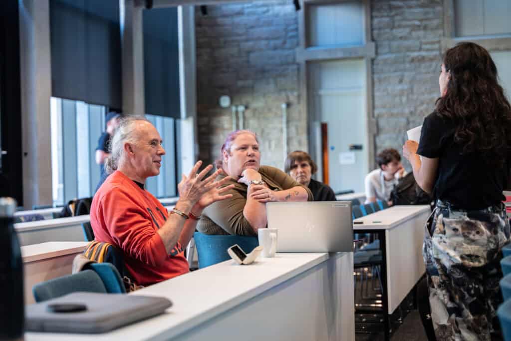 Three people sit at desks in a classroom, engaged in conversation with a standing woman. One man gestures with his hands, while others listen. A laptop and mug are on the desk in front of them.