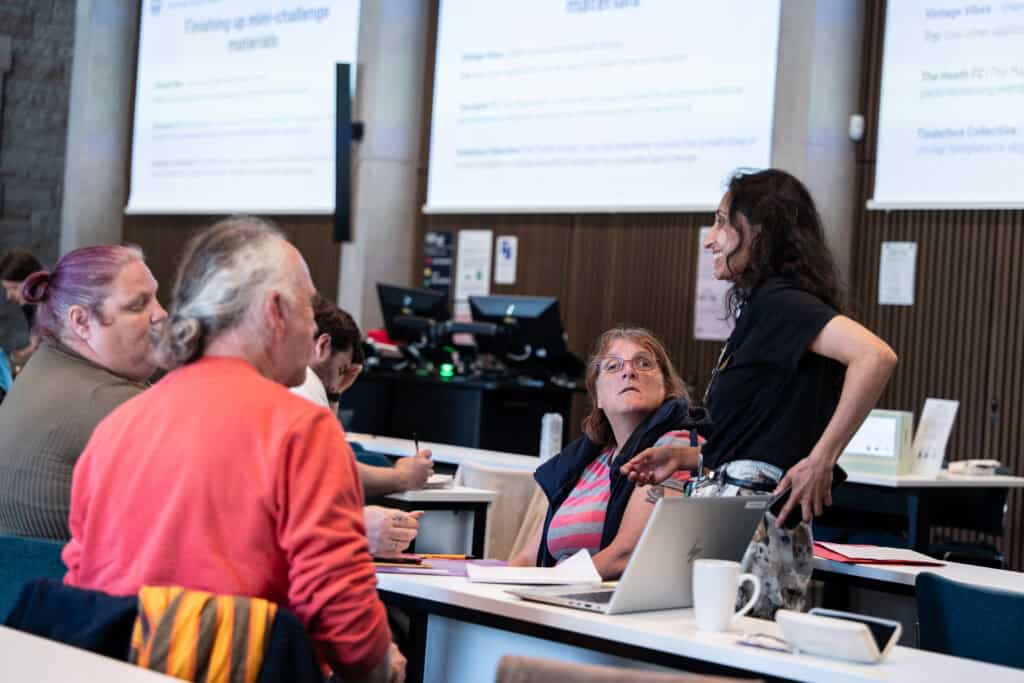 A group of adults sit at desks in a classroom, engaged in discussion. One woman stands, speaking to seated participants. Presentation slides are projected on screens at the front of the room. Laptops and notebooks are on the tables.