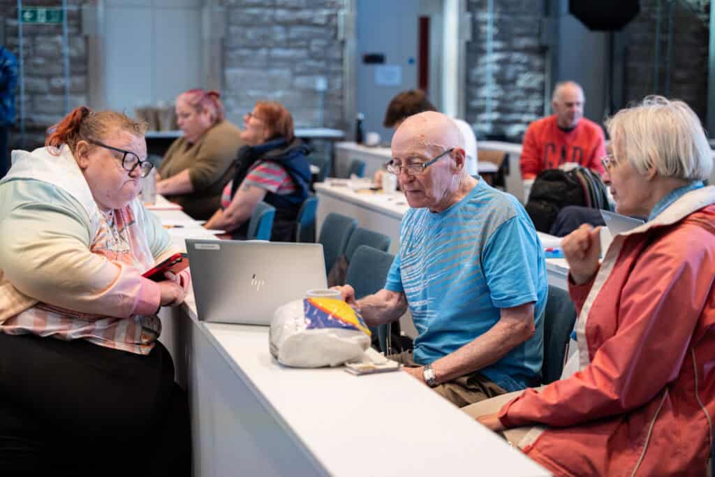 Several adults sit in a classroom, some talking and others using laptops. In the foreground, an older man and woman converse, while a woman beside them looks at her laptop and mobile. The setting appears casual and collaborative.