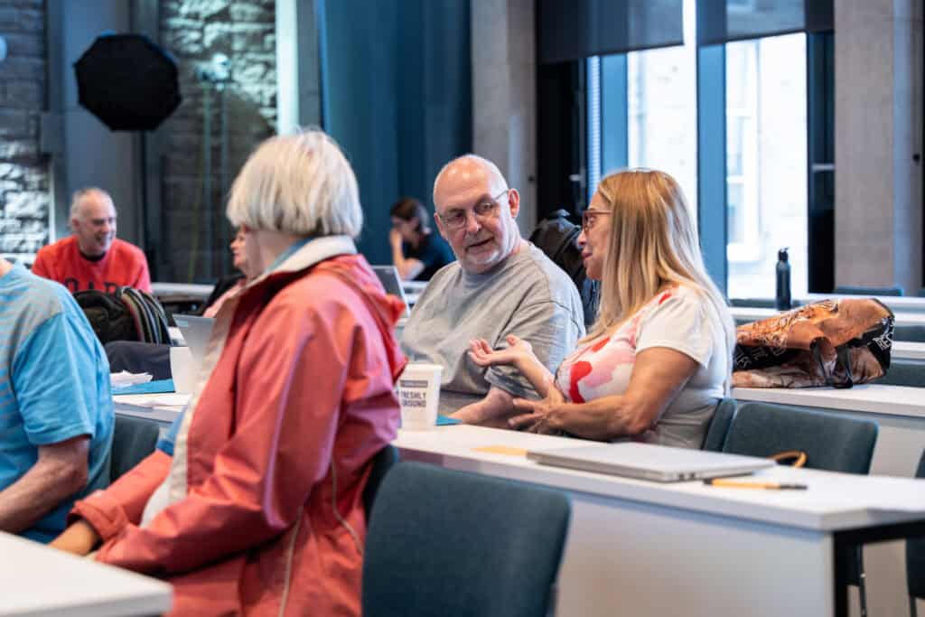 Several older adults sit at desks in a classroom. Two people in the centre have a conversation. Notebooks, laptops, and bags are visible on the desks. Sunlight comes through the large windows in the background.