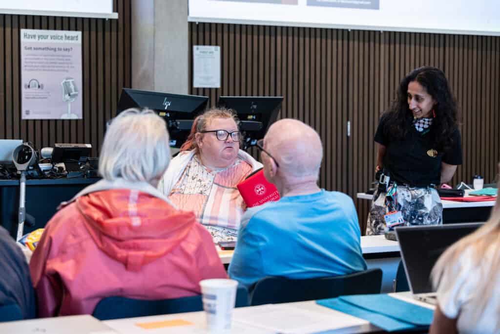 A group of people sit at tables in a classroom, engaged in discussion. One woman stands smiling near a desk, whilst others face her, listening attentively. Computers and a cup are visible in the background.