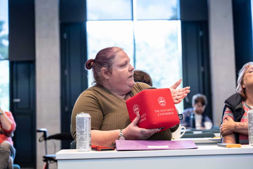 A person with red hair speaks while holding a red cube with “The University of Edinburgh” printed on it, sitting at a desk with a drinks can and purple folder in a modern classroom.