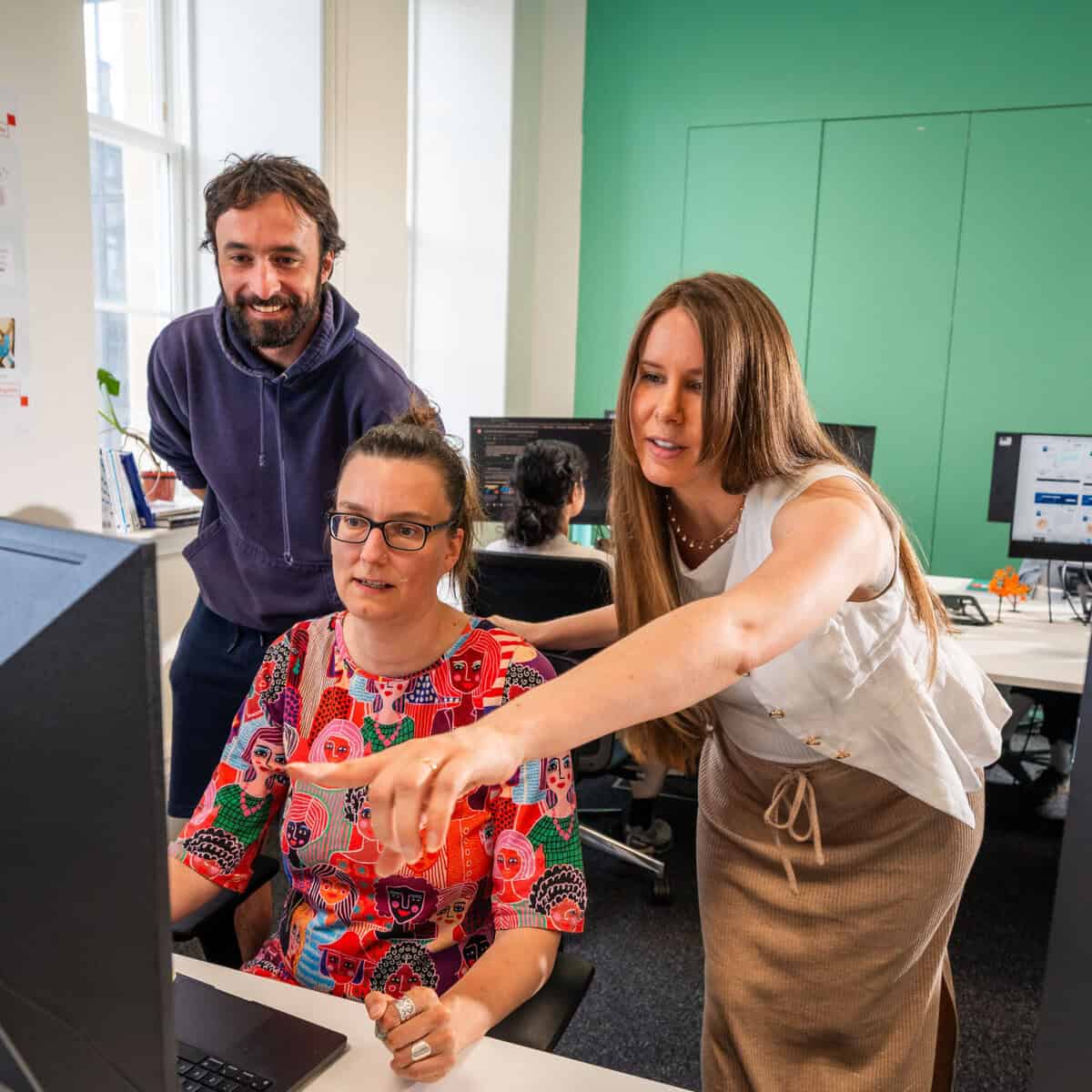 Three people collaborate at a computer in a bright office. A woman sitting wears glasses, while another woman stands pointing at the screen. A man stands behind them, smiling. Monitors and posters are visible in the background.