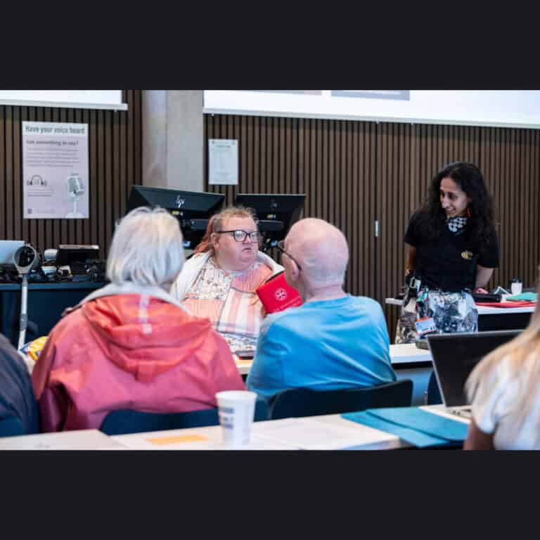 A group of people sit at a table in a meeting room whilst a woman stands nearby, listening. Computer monitors and informational posters are visible in the background.