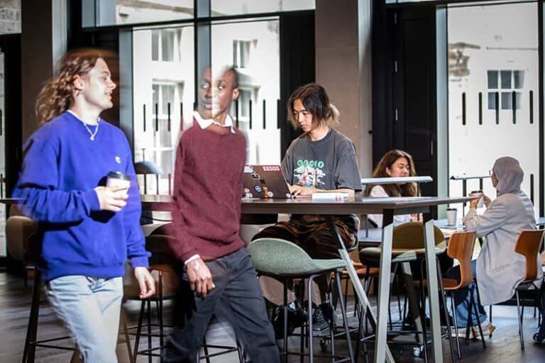 Several people gather in a modern café; two walk past each other, others sit at tables working on laptops or talking. The space is bright with large windows in the background.