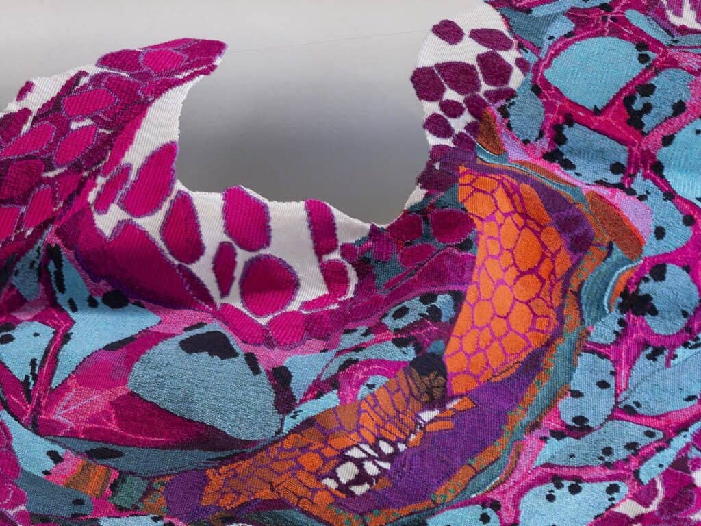 Colourful, textured abstract fabric with bright magenta, blue, orange, and black patterns. The edges are irregular and the surface appears wavy, giving a dynamic, sculptural effect.