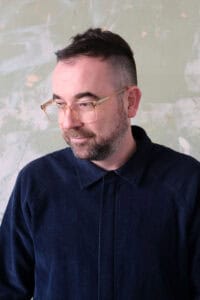 A man with short dark hair, glasses, and a beard wears a navy corduroy shirt and looks slightly downwards against a textured, light-coloured background.