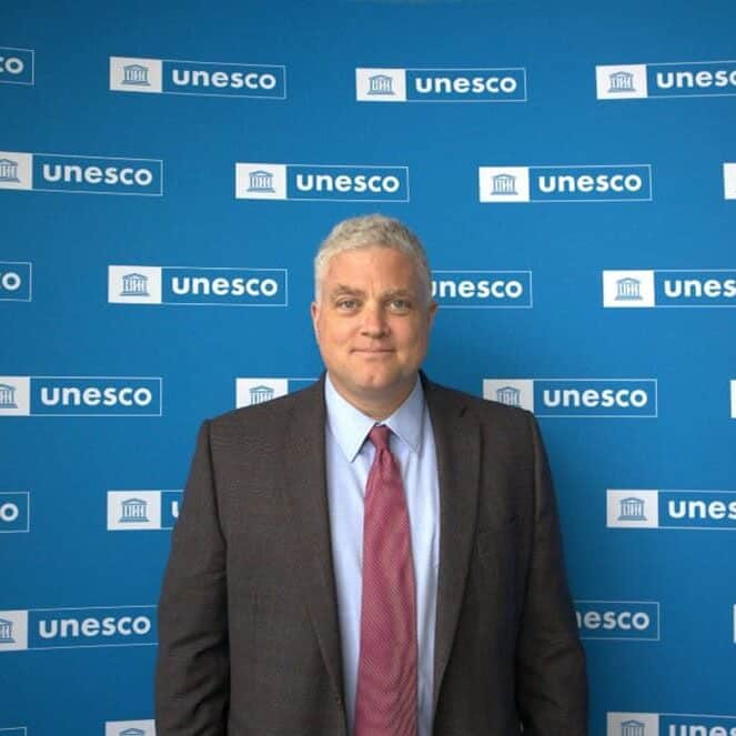 A man in a suit and red tie stands in front of a blue backdrop featuring multiple white UNESCO logos.