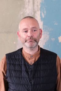 A man with a close-cropped beard and short hair, wearing a brown shirt and black quilted gilet, stands against a textured wall painted in faded green and blue.