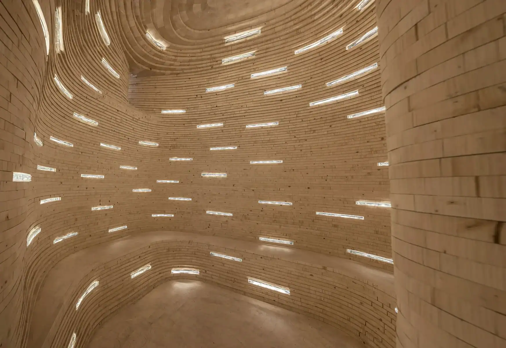 Curved walls made of tan bricks, with thin, horizontal slit windows allowing light in at regular intervals, creating a pattern on the wavy interior surfaces.