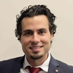 A smiling man with curly dark hair, a trimmed goatee, and dimples, wearing a dark suit, white shirt, and red tie, standing in front of a plain light background.