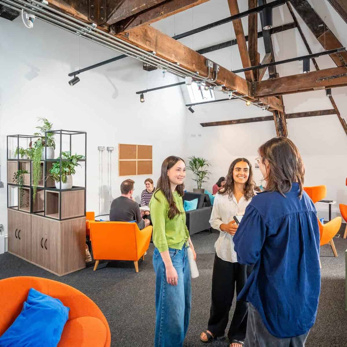 People talking and relaxing in a modern office lounge with orange chairs, green door, exposed beams, and plants. Several groups are engaged in conversation, creating a friendly and collaborative atmosphere.
