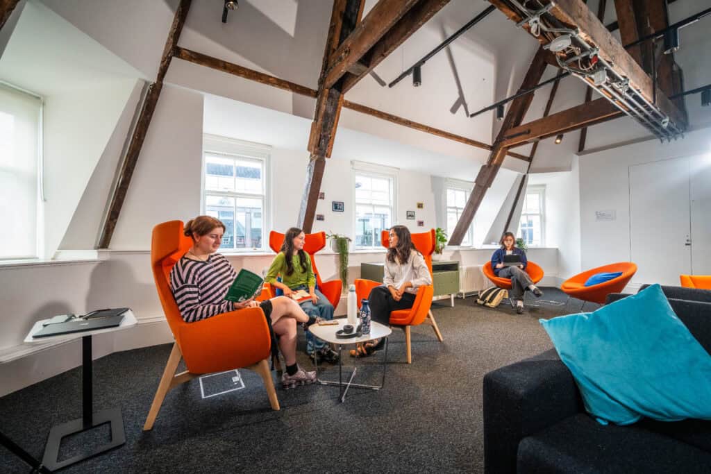 Four people sit on orange chairs in a modern office with exposed beams, talking and working. Large windows let in natural light. A sofa with blue cushions is visible in the foreground.