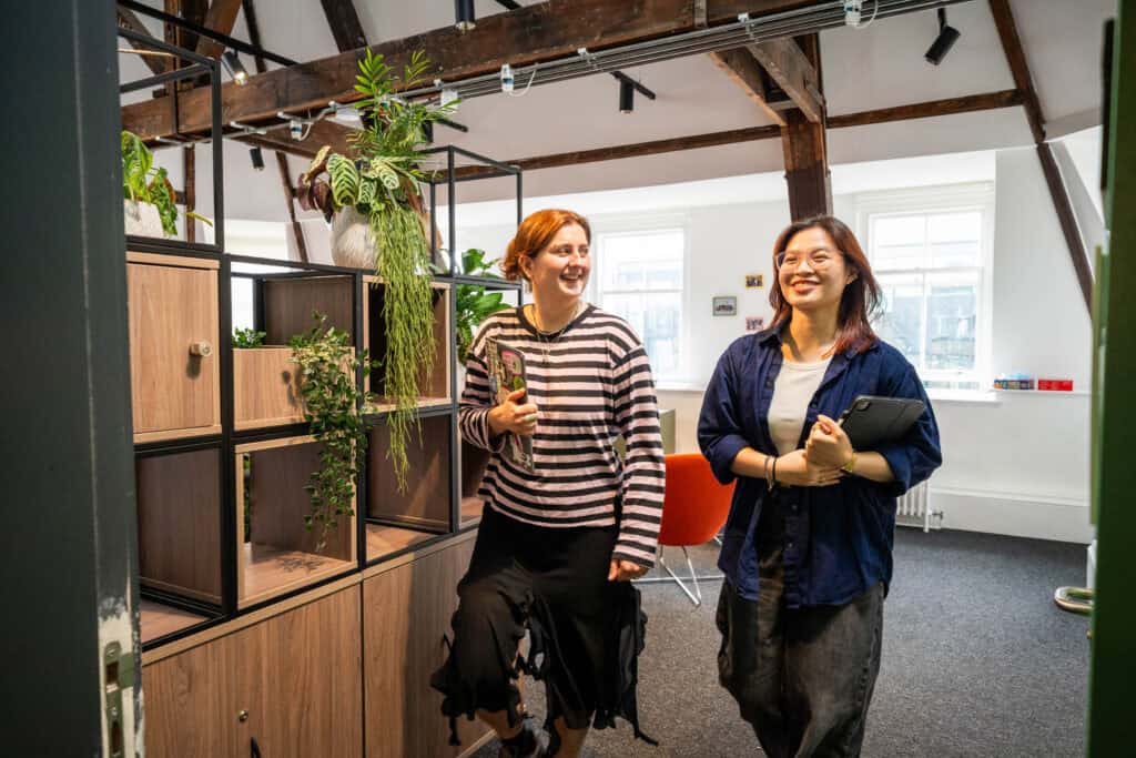 Two people smile and walk into a bright office space with exposed beams, wooden shelves, and green plants. One holds a drink, the other carries a laptop.